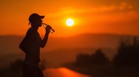 Silhouette of person drinking water from bottle against dramatic orange sunset sky with bright sun and mountain backdrop.