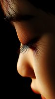 Close-up profile view of long eyelashes and delicate facial features illuminated dramatically against dark background, showing subtle skin texture and natural beauty.