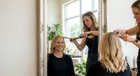 Young Caucasian woman smiling in mirror while hairstylist curls her blonde hair in a bright modern salon setting.
