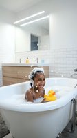 Happy African American child enjoying bubble bath with rubber duck toy in modern white bathroom with wooden vanity and LED mirror lighting.
