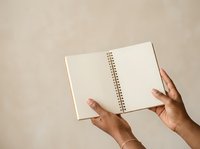 Open spiral notebook with blank pages held by hands against neutral beige background, suitable for stationery, education, planning, and office concepts.