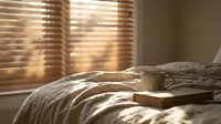 Cozy bedroom scene with open book on unmade bed, warm sunlight streaming through wooden blinds creating peaceful morning atmosphere for relaxation.