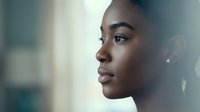 Young African American woman in profile with natural makeup and earrings looking thoughtfully into distance with soft lighting and blurred background.