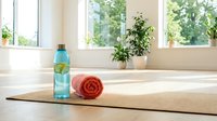 Wellness setup with infused water bottle, rolled towel, and potted plants on yoga mat near bright windows for fitness and relaxation routines.
