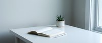 Minimalist workspace with open book, notebook, and potted succulent plant on white desk near bright window for creative work and study.