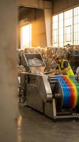 Industrial worker operating printing machinery with colorful paper rolls in manufacturing facility with natural sunlight streaming through windows.