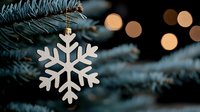 White snowflake ornament hanging on pine tree branch with soft bokeh lights in background creating magical winter holiday atmosphere.