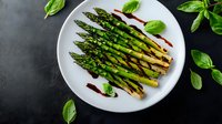 Grilled green asparagus spears on white plate garnished with fresh basil leaves and balsamic reduction, served on dark background. Food photography.