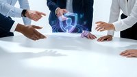 Business professionals in formal attire gathered around a white table with glowing digital shield icon, representing cybersecurity, data protection, and secure network collaboration.