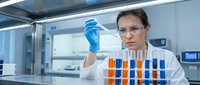 Female scientist in protective eyewear examining blue liquid sample with pipette in modern laboratory. Research, testing, analysis, chemistry experiments.