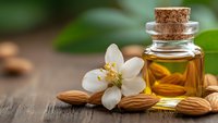 Essential oil bottle with cork stopper, white jasmine flower and almonds on wooden surface for aromatherapy and natural wellness treatments.