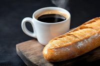 Fresh crusty baguette on rustic wooden board with white cup of hot black coffee against dark background, morning breakfast concept.