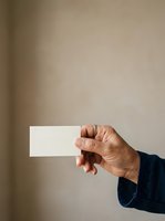 Adult male hand with medium skin tone holding blank white business card or paper against neutral beige wall background for corporate branding and identity mockup purposes.