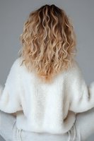 Back view of blonde wavy hair with balayage highlights on young woman wearing cozy white knit sweater against grey background. Modern hairstyling concept.