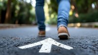 White arrow painted on asphalt path with blurred walking feet in blue jeans and sneakers, shallow depth of field view showing direction and movement forward.