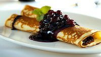 Thin golden crepe rolled and served on white plate with dark berry sauce and fresh mint garnish, close-up view showing delicate texture and glossy topping.