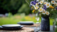 Fresh wildflower bouquet with daisies and purple blooms in navy textured vase on rustic outdoor table setting with woven placemats and dark plates against blurred garden.