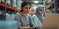 Young diverse female professionals working together at laptop in modern warehouse office space with industrial storage shelves in background.