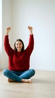 Young woman in red sweater celebrating success with raised arms sitting cross-legged on wooden floor against white wall expressing joy and achievement.