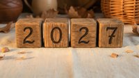 Wooden blocks spelling 2027 on a rustic linen surface with autumn leaves and a wicker basket, evoking a warm, cozy new year concept.