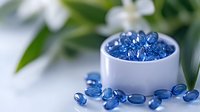 Close-up of blue gel capsules spilling from white container with green foliage background, suitable for pharmaceutical, wellness and healthcare marketing materials.