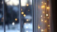 Glowing fairy lights create magical bokeh effect against window at dusk, with soft winter snow scene outside, creating cozy atmospheric holiday mood.