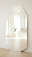 Organic-shaped mirror reflecting minimalist interior with natural light, potted plant, wooden stool, and ceramic vessel. Suitable for home decor, interior design, and lifestyle content.