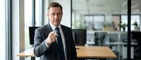 Professional businessman in suit pointing at camera in modern office workspace with glass walls and wooden desk.