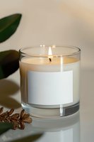 Aromatic candle in glass container with blank label, burning flame creates cozy atmosphere. Green leaves and dried pine cone add natural spa-like ambiance.