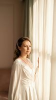 Young Caucasian bride in elegant white wedding dress with lace details looking thoughtfully out window with soft natural light streaming through curtains.