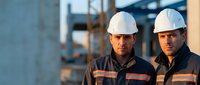 Two professional construction workers in white hard hats and safety uniforms standing at industrial building site during golden hour lighting.