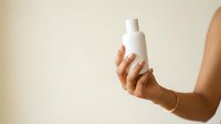 Woman with medium skin tone holding white cosmetic bottle against neutral background for skincare product promotion.