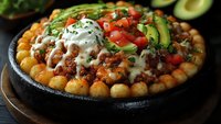 Loaded tater tots topped with seasoned ground beef, melted cheese, sour cream, diced tomatoes, avocado and fresh herbs on cast iron skillet, comfort food appetizer.