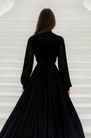 Young woman in elegant black pleated maxi dress with long sleeves shown from behind, standing against white background. Classic fashion portrait composition.