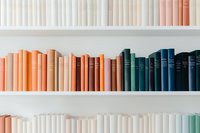 Books arranged by color on white shelves creating ombre effect from white through peach and orange to deep blue tones, modern minimalist library organization.