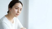Young Asian woman in white shirt sitting thoughtfully by window with natural light, contemplative mood for wellness and lifestyle content.