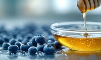 Fresh ripe blueberries scattered on blue surface with golden honey dripping from wooden dipper into glass bowl, macro food photography.