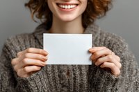 Smiling woman with curly hair in cozy sweater holding blank white card for your message against gray background.