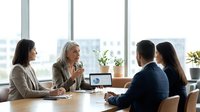 Mature female executive with blonde hair leading business meeting with diverse team in modern office with city skyline view, discussing strategy and collaboration.