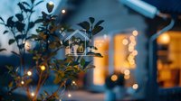 Digital house icon with glowing lines over blurred evening garden background with bokeh lights and plant silhouettes, concept of smart home technology.
