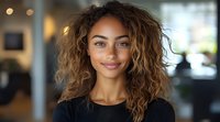 Young mixed race woman with curly highlighted hair and natural makeup smiling warmly at camera. Professional headshot style portrait with soft bokeh background.