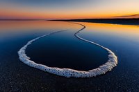Curved line of salt deposits along dark shore creates dramatic contrast against calm water surface reflecting vibrant sunset sky in peaceful natural landscape.