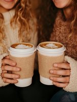 Two takeaway coffee cups with latte art held by women in cozy sweaters. Warm tones and intimate cropped composition capture friendship and coffee culture moment.