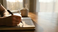 Person writing in notebook with pen at wooden desk near window with warm sunlight, coffee cup beside, suitable for productivity, education, journaling, and creative work concepts.