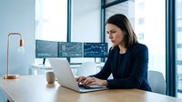 Professional businesswoman working on laptop in modern office with multiple monitors and city view windows for corporate presentations.