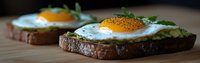Open-faced sandwich with sunny side up eggs and mashed avocado on dark toasted bread, garnished with fresh herbs and black pepper, served on wooden surface.