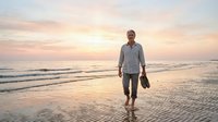 Middle-aged man with light skin tone walking barefoot on sandy beach at sunset, carrying shoes in hand, wearing casual button-up shirt and shorts. Serene coastal scene for lifestyle and wellness content.