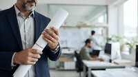 Mature male professional with beard wearing dark blazer and light shirt holding rolled white blueprint document in modern office environment with colleagues working at desks in background.