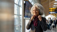 Mature African American businesswoman with curly gray hair adjusting scarf at modern airport terminal with departure boards and travelers in background.