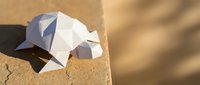 White geometric low poly paper turtle sculpture resting on a sandy stone surface in warm sunlight with soft shadows.